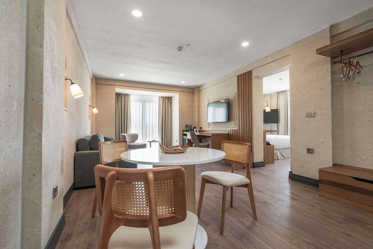 Ein modernes Hotelzimmer mit einem runden Esstisch, Holzstühlen, einer Couch, einem Fernseher und einem Holzschrank.