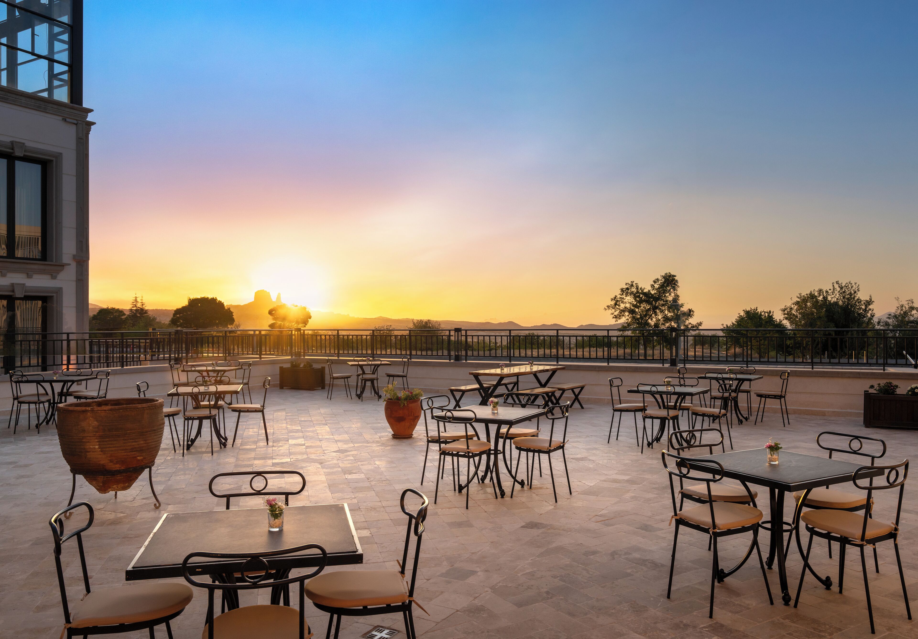 Un patio exterior con mesas y sillas, una maceta grande y vistas al atardecer.