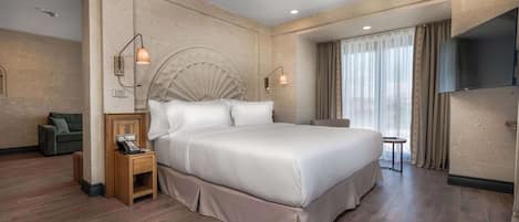 Amplia habitación de hotel con una cama grande, una mesita de noche de madera, una pequeña mesa, un sofá y un televisor.