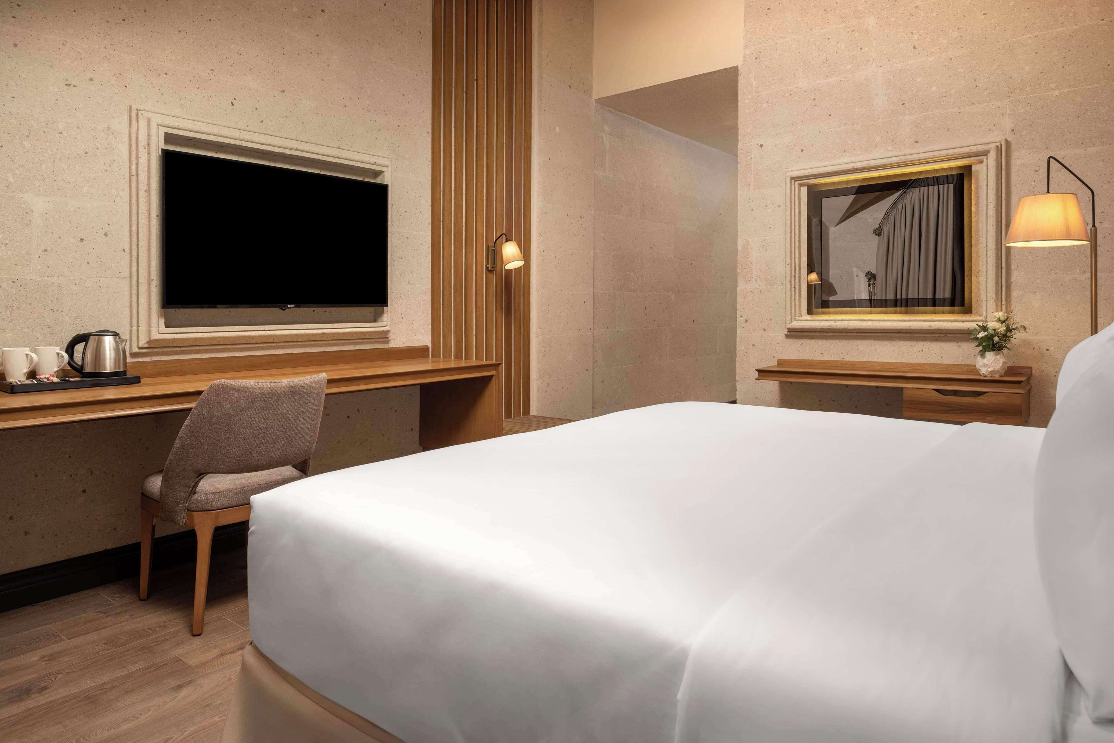 A hotel room with a bed, a desk with a chair, a television, a lamp, and a window with curtains.