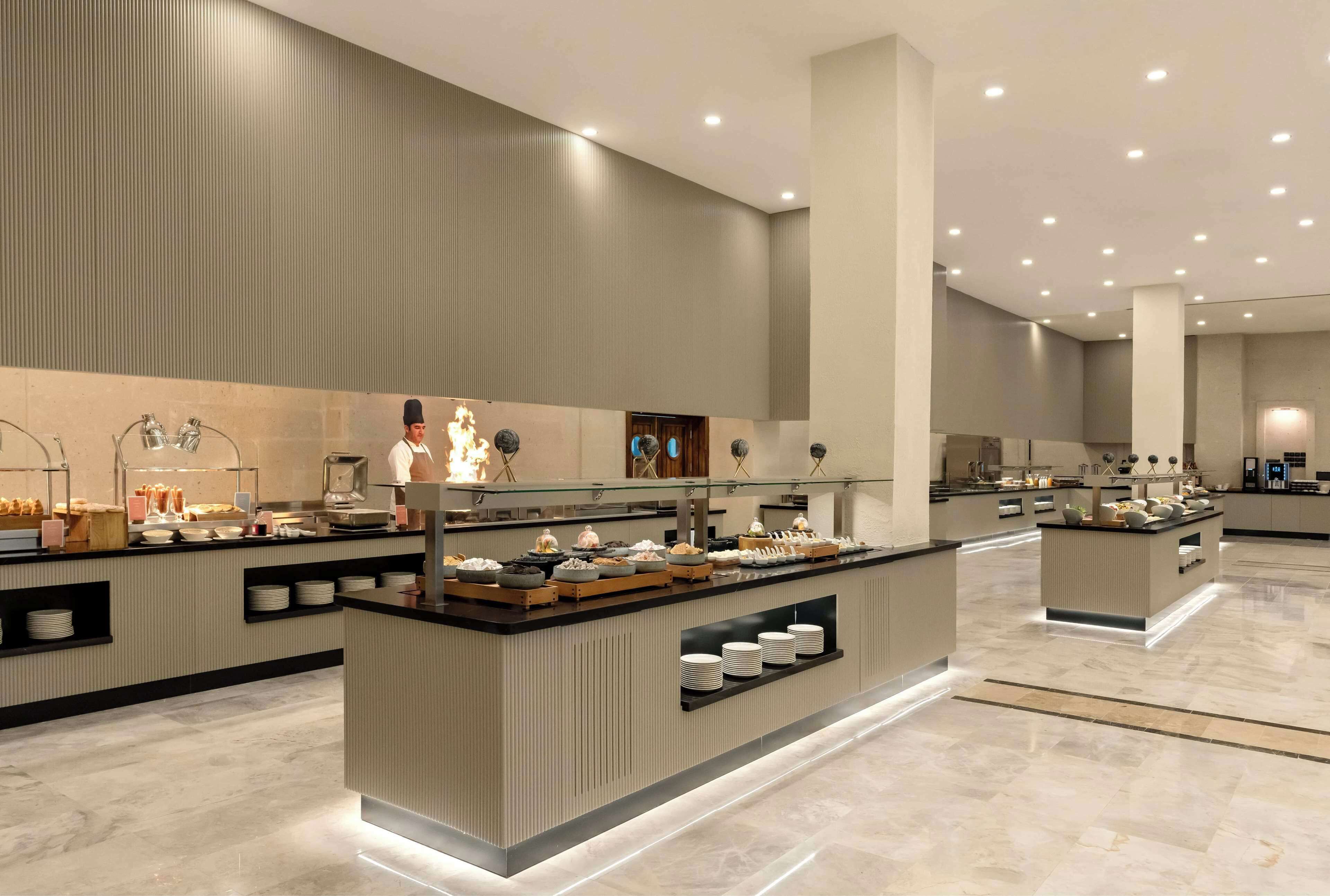 Un moderno vestíbulo de hotel con un diseño espacioso, un mostrador de recepción central y una zona de buffet con diversas opciones de comida.