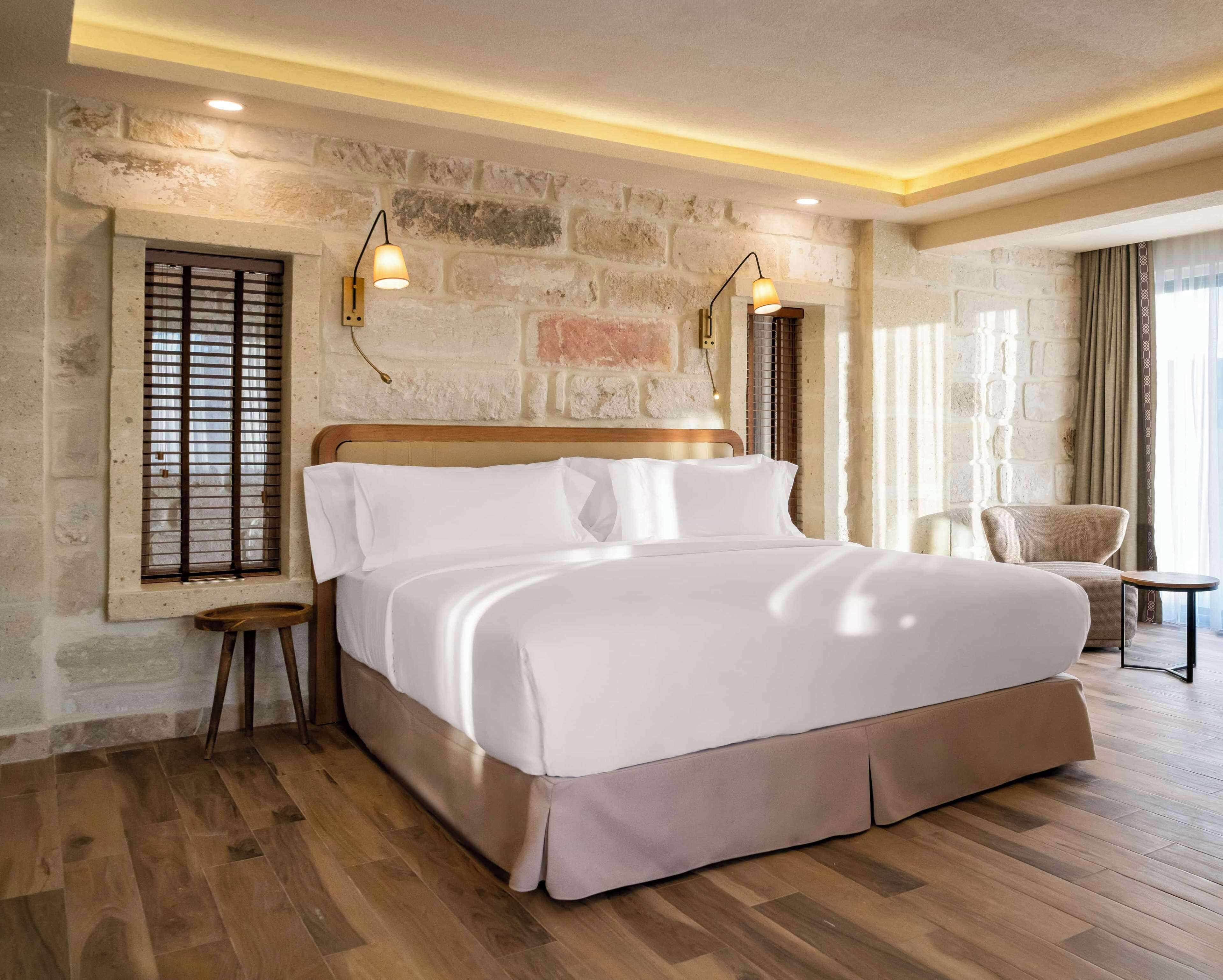 A spacious bedroom with a large bed, wooden flooring, stone wall, and a seating area with a chair and table.