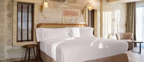 A spacious bedroom with a large bed, wooden flooring, stone wall, and a seating area with a chair and table.