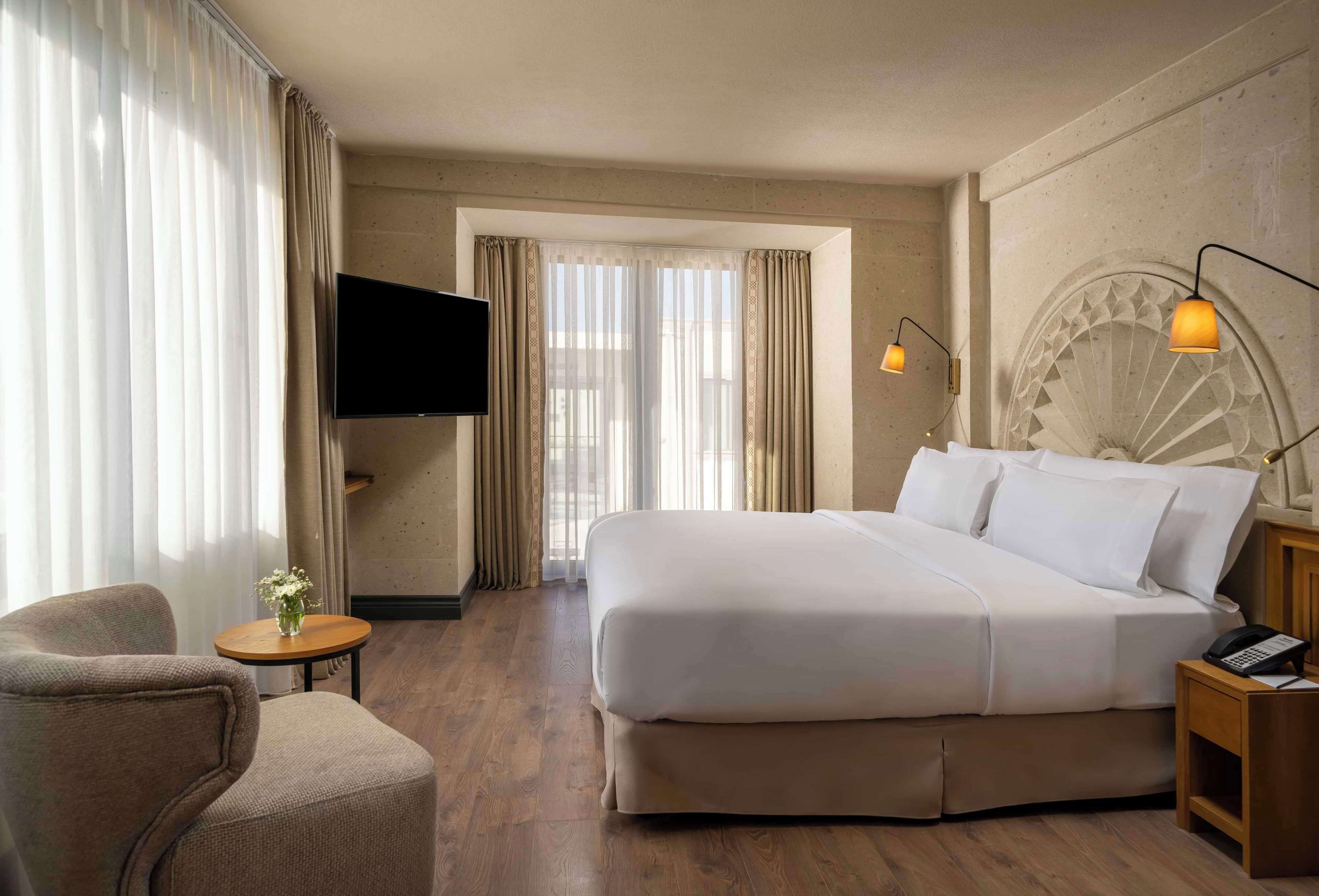Ein Hotelzimmer mit einem großen Bett, einem Sessel, einem kleinen Tisch, einem Fernseher und einer Lampe.