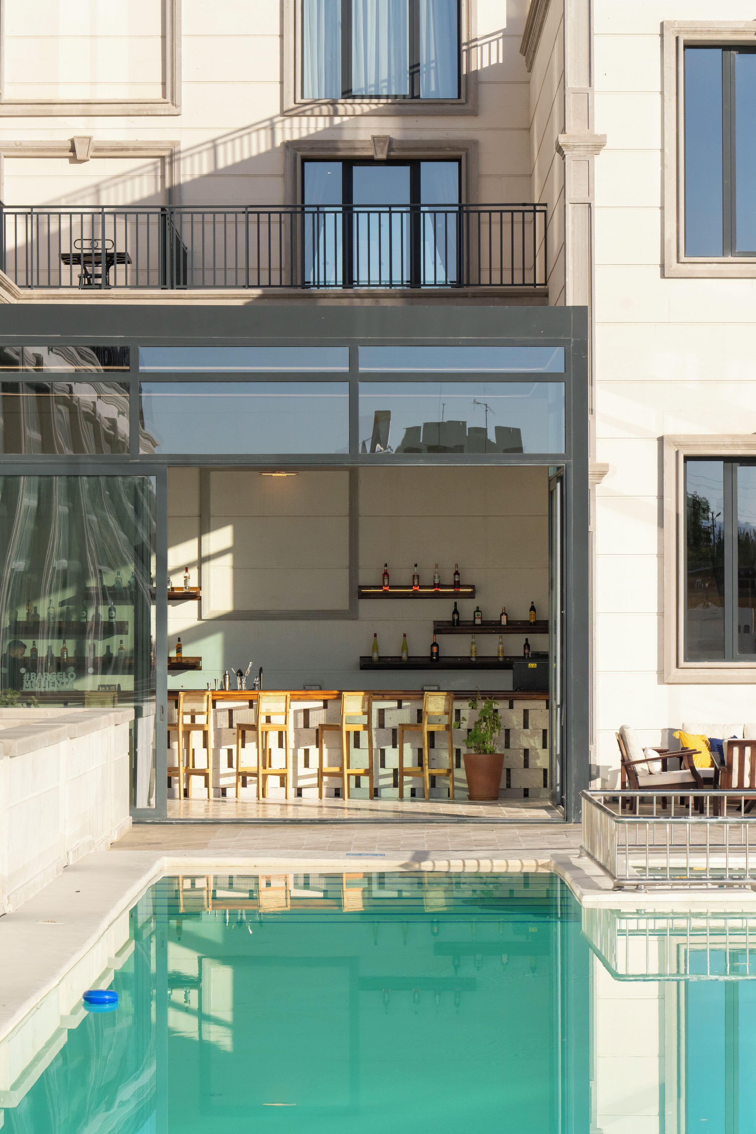 Un edificio moderno de dos pisos con un área de piscina cubierta de vidrio, un balcón con banca y una cocina con estantes llenos de botellas.