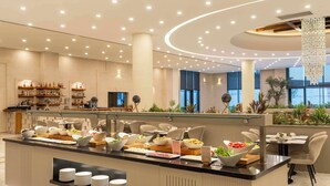 Um hall de hotel moderno com área de buffet, espaços para sentar e uma grande janela com vista para a cidade.