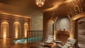 Uma área de piscina interior luxuosa, com piso de mármore, paredes de pedra e duas cadeiras brancas acolchoadas.