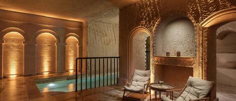 Un área de piscina interior lujosa con suelo de mármol, paredes de piedra y dos sillones blancos acolchados.