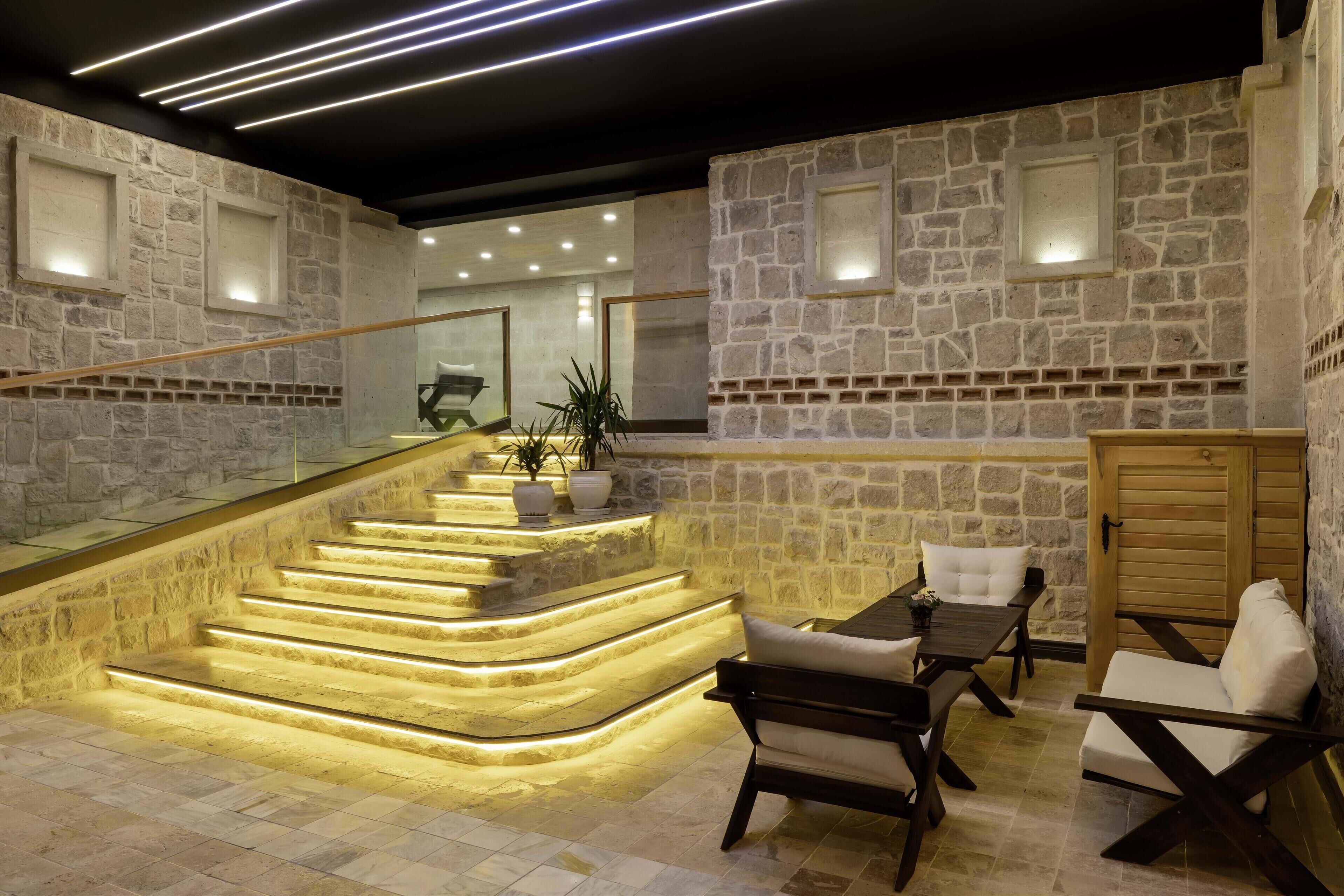 Un interior moderno con paredes de piedra, una escalera con iluminación integrada y una zona de estar con mesa y sillas.