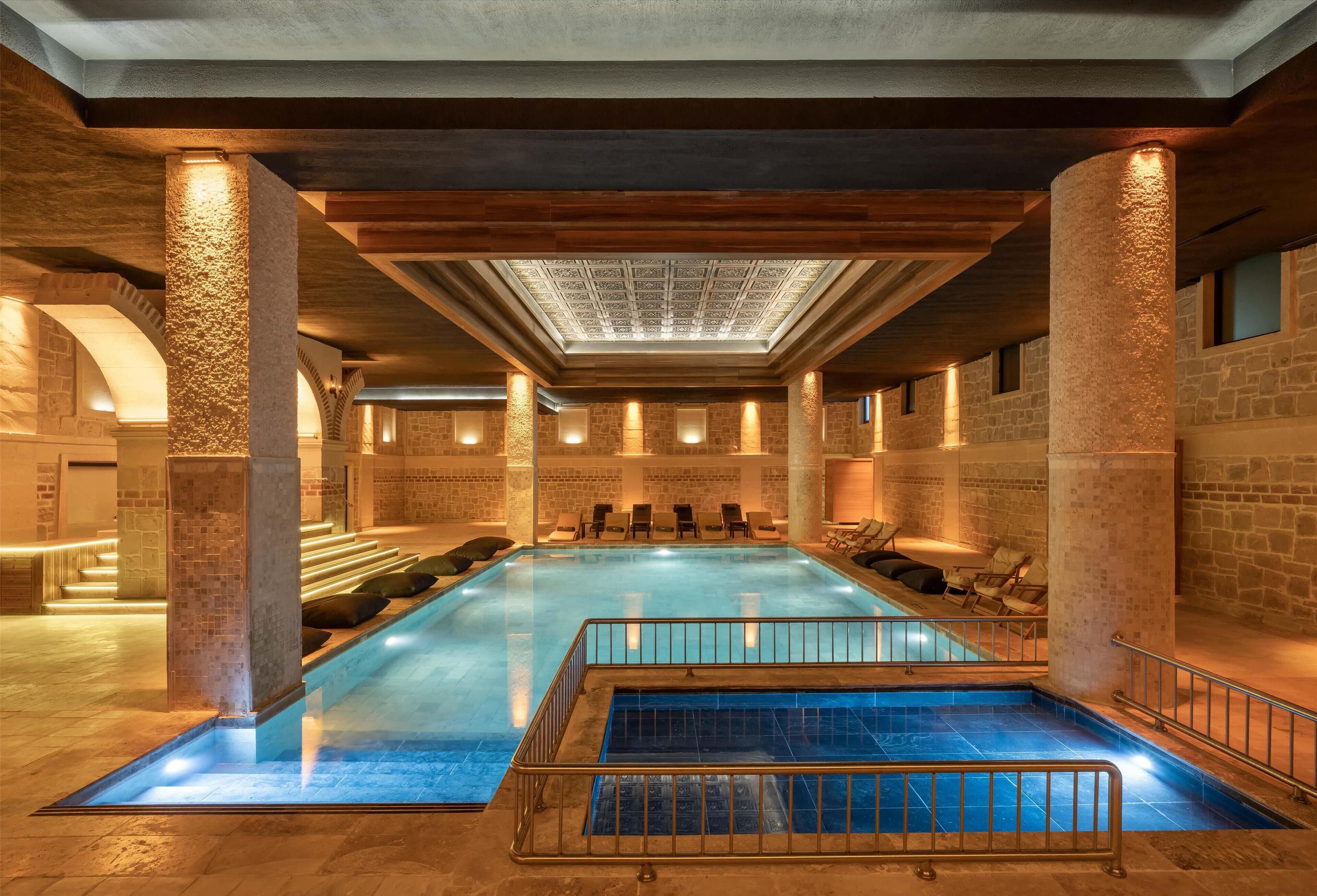 Una amplia piscina interior con techo de tragaluces, paredes de piedra y varias zonas de descanso.