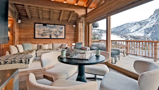 L’intérieur d’un chalet en bois chaleureux et accueillant, avec un canapé, des fauteuils et une table. Un tableau est accroché au mur, et on peut admirer, par la fenêtre, des montagnes enneigées.