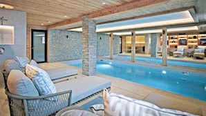 Un espace piscine intérieur moderne, avec des murs en pierre, des poutres en bois et des sièges disposés au bord de la piscine.