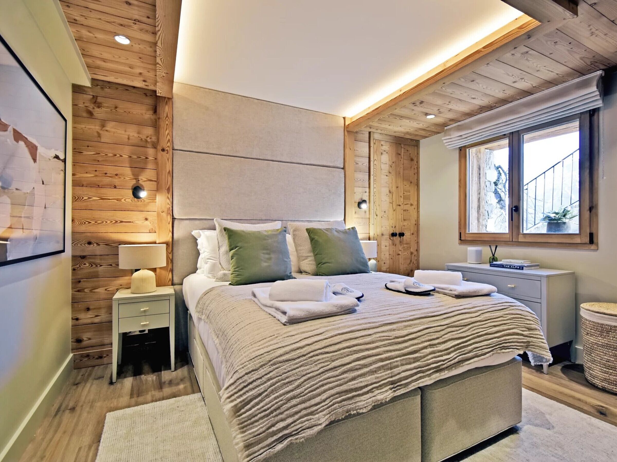A modern bedroom with a wooden ceiling, a large bed with multiple pillows, a bedside table with a lamp, and a window with a view.