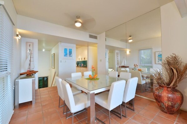 A dining area with a glass table and white chairs, a large vase with dried plants, and a kitchen in the background. - Port Ridge; The Hill Penthouse (Port Douglas)