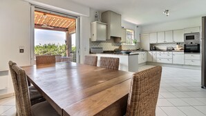 A modern kitchen with white cabinets, a wooden dining table, wicker chairs, and a view of greenery outside. - Beautiful villa with stunning view nestled in countryside near Valbonne village  (OPIO)