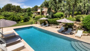 A rectangular swimming pool with clear blue water, surrounded by a stone deck. There are lounge chairs and umbrellas, and a house is visible in the background. - Beautiful villa with stunning view nestled in countryside near Valbonne village  (OPIO)