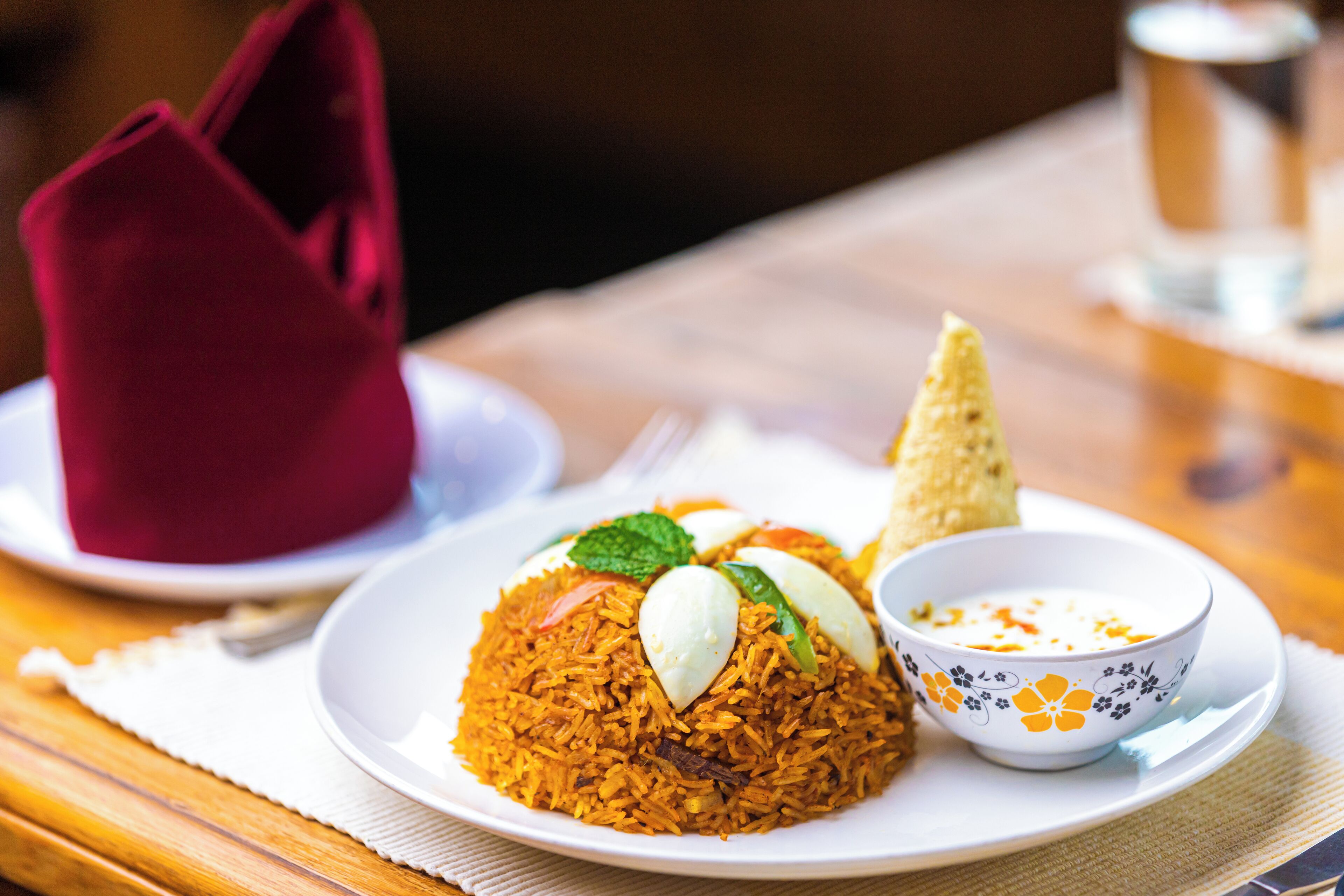 A plate of biryani with boiled eggs and a side of raita, served on a white plate with a floral pattern.