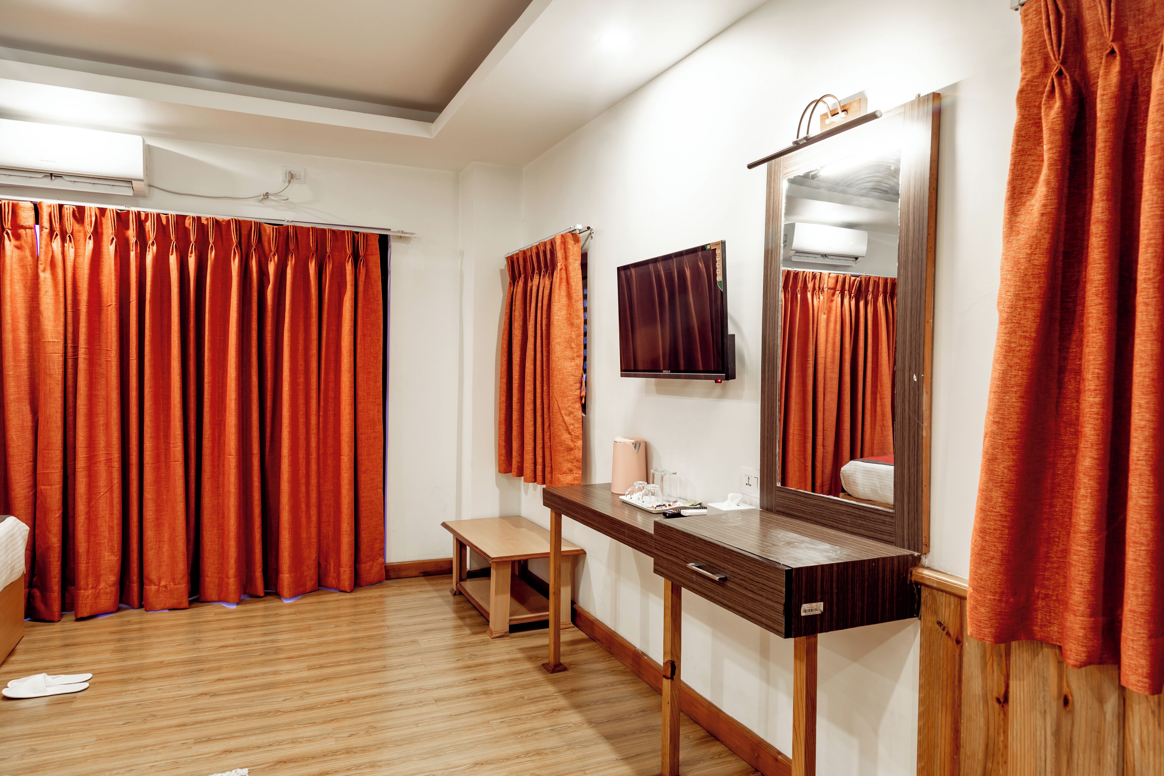 A hotel room with a wooden floor, a wooden desk, a mirror, and orange curtains.