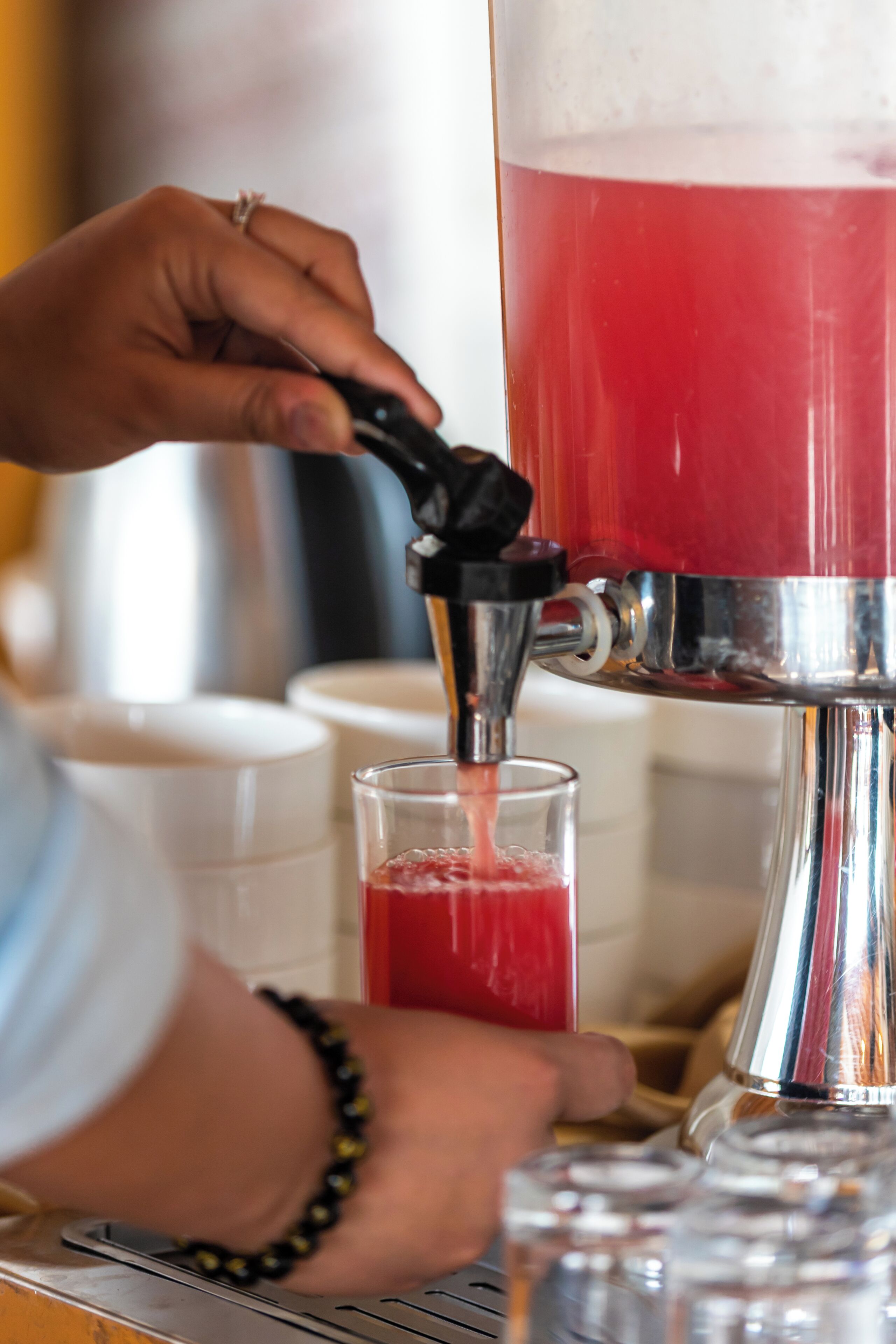 A person pouring a red beverage from a metallic dispenser into a clear glass.
