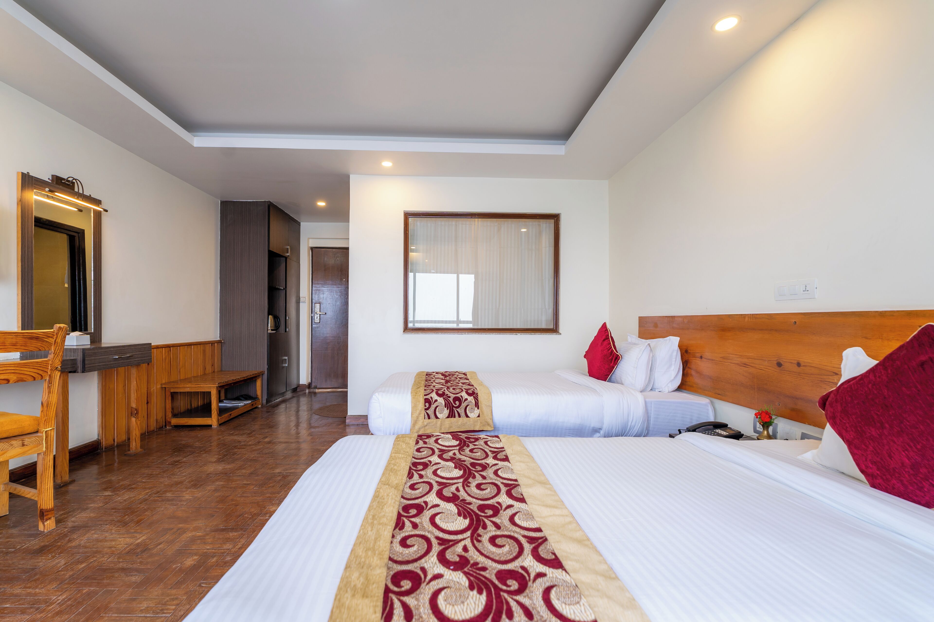 A hotel room with two beds, a wooden desk, a mirror, and a large window.