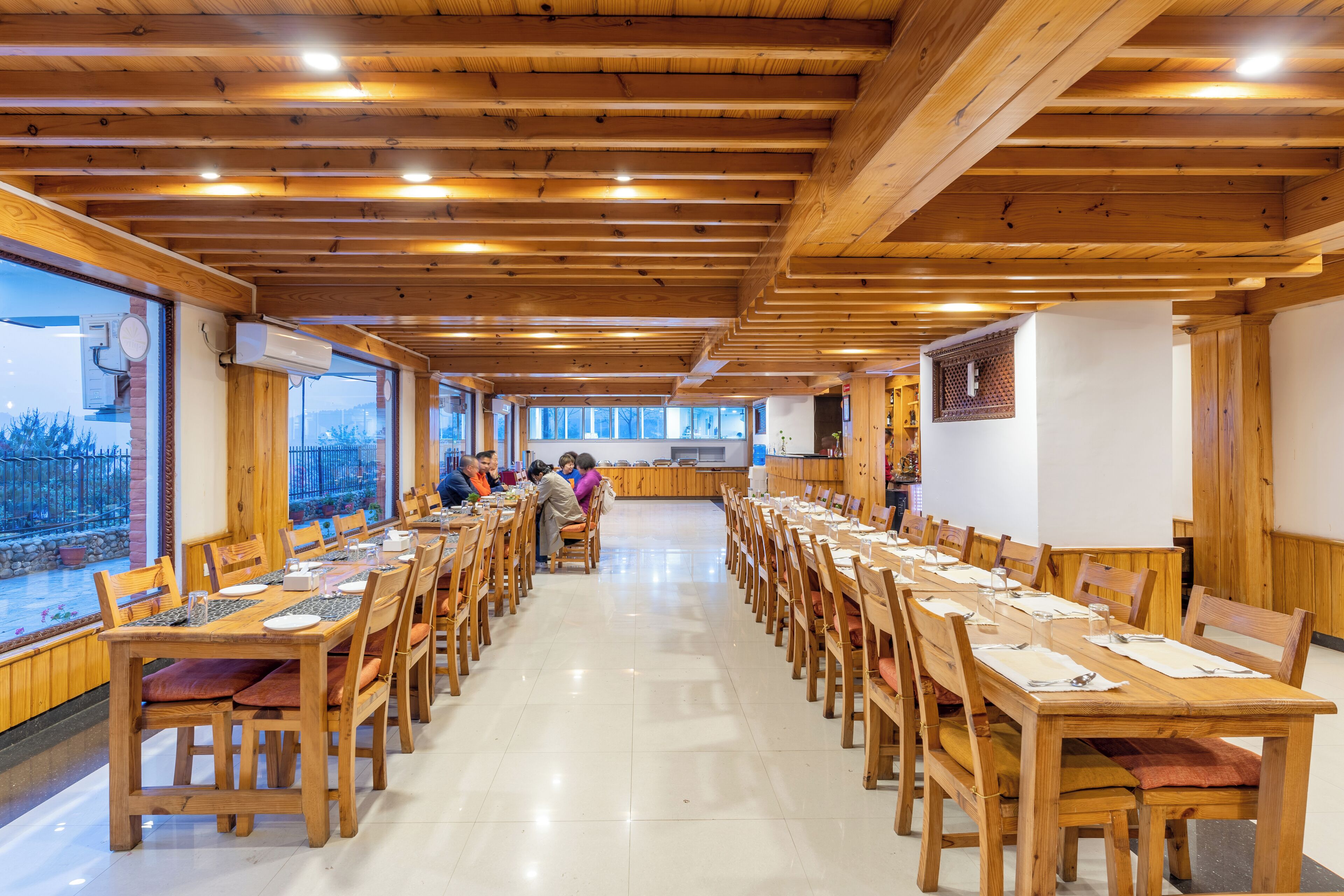 A long wooden dining table with chairs, surrounded by large windows and wooden beams on the ceiling.