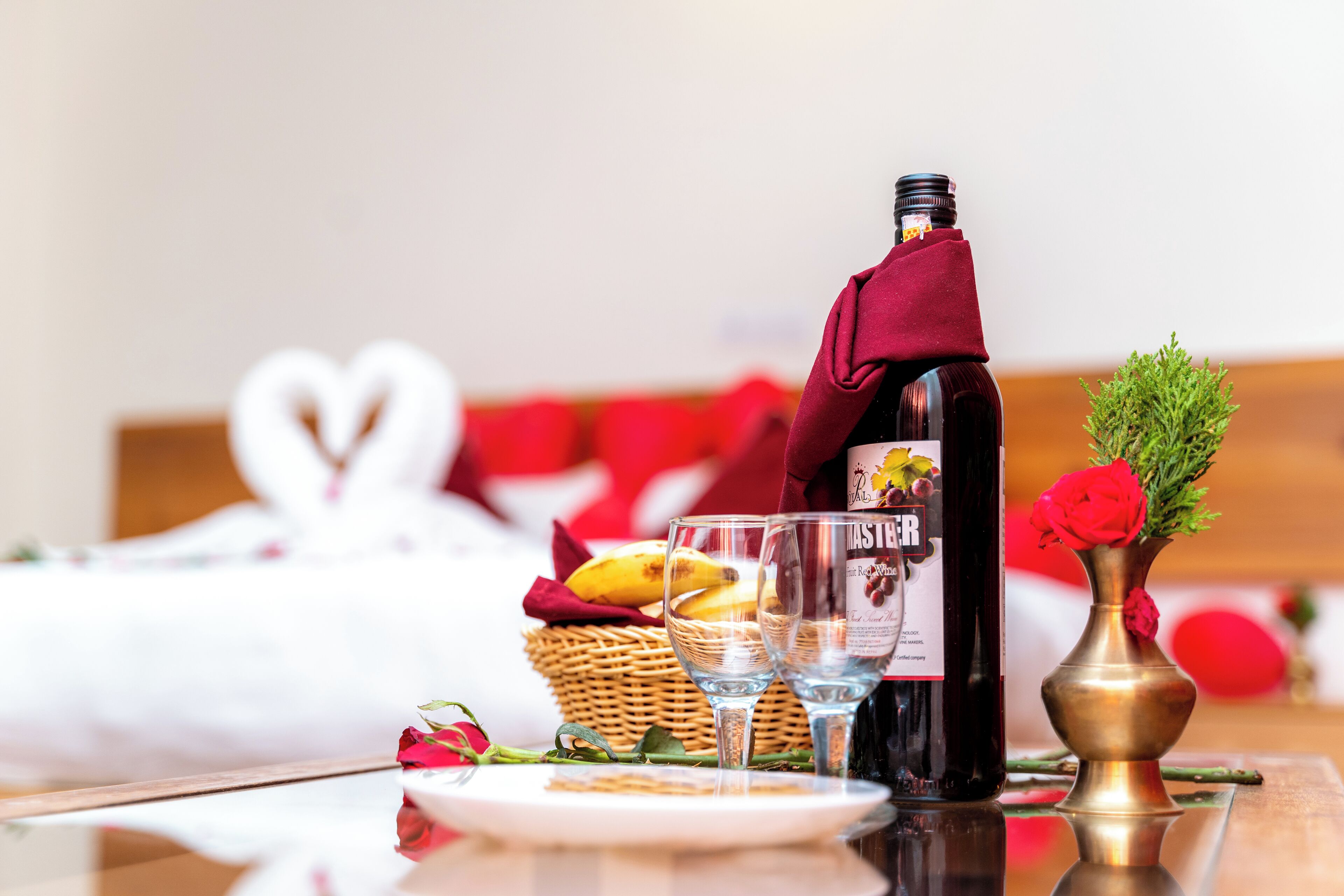 A table with a bottle of wine, two glasses, a basket with bananas, a rose, and a vase with a red rose.