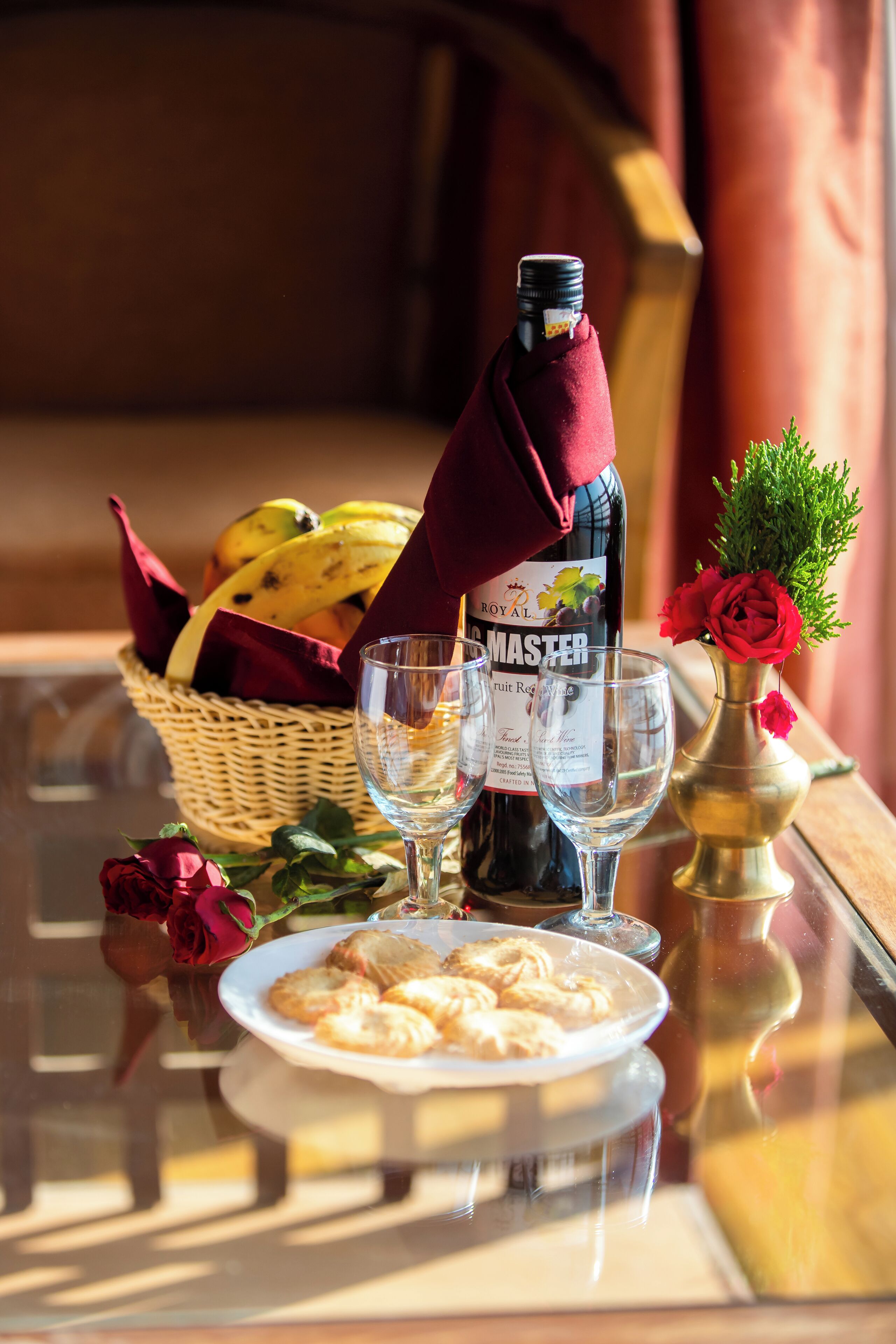Una tavola apparecchiata con una bottiglia di vino, bicchieri, un cesto di frutta, un piatto di pasticcini e un vaso con un fiore.