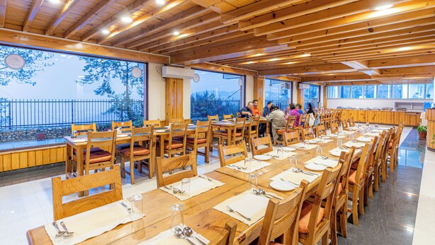 Un vaste espace dédié à la salle à manger, avec des tables et des chaises en bois, de grandes fenêtres offrant une vue sur l’extérieur et un design moderne au niveau du plafond.