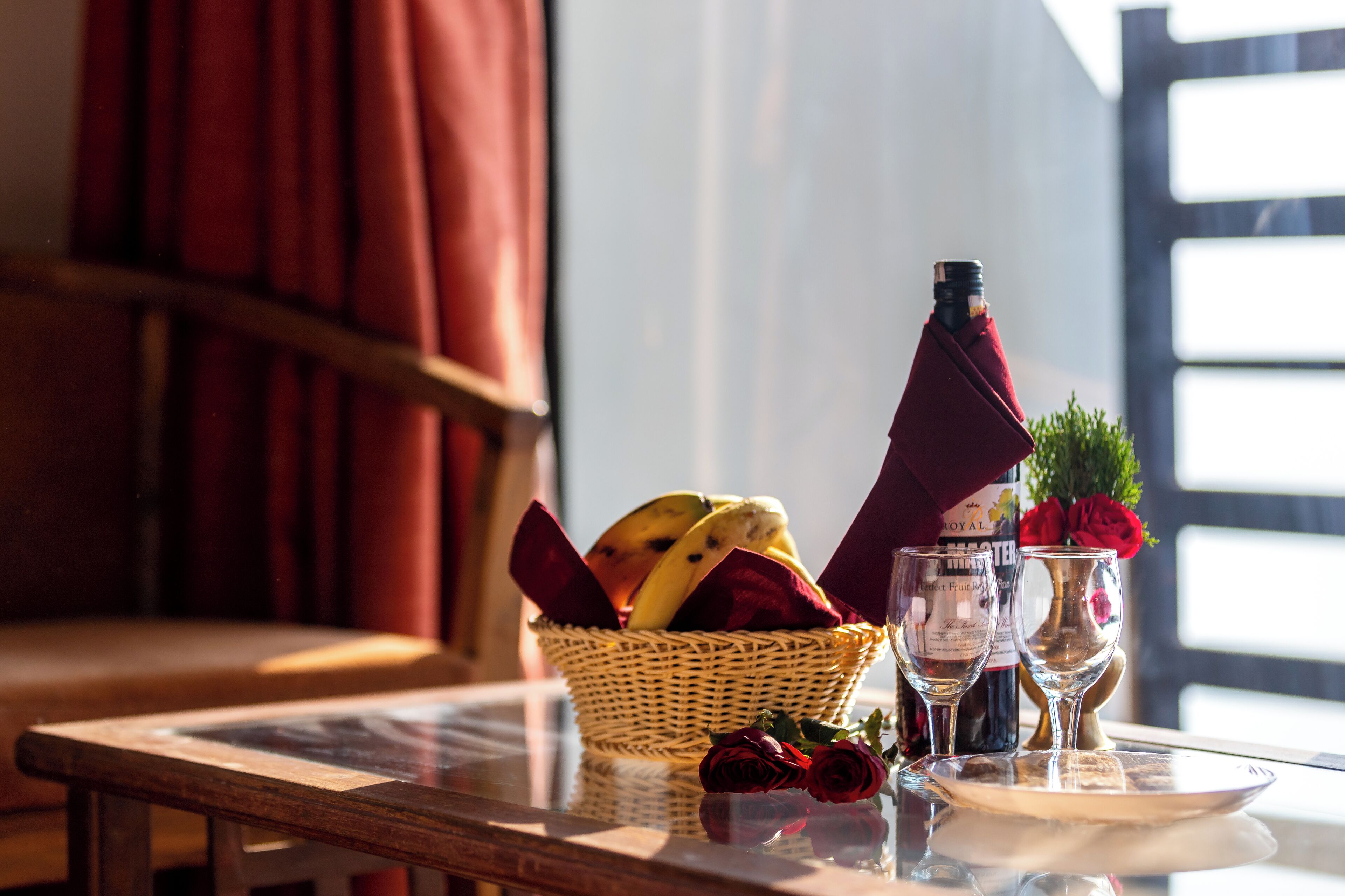 Een tafel met een rieten mand vol fruit, een wijnfles, twee wijnglazen en een vaas met een rode roos.
