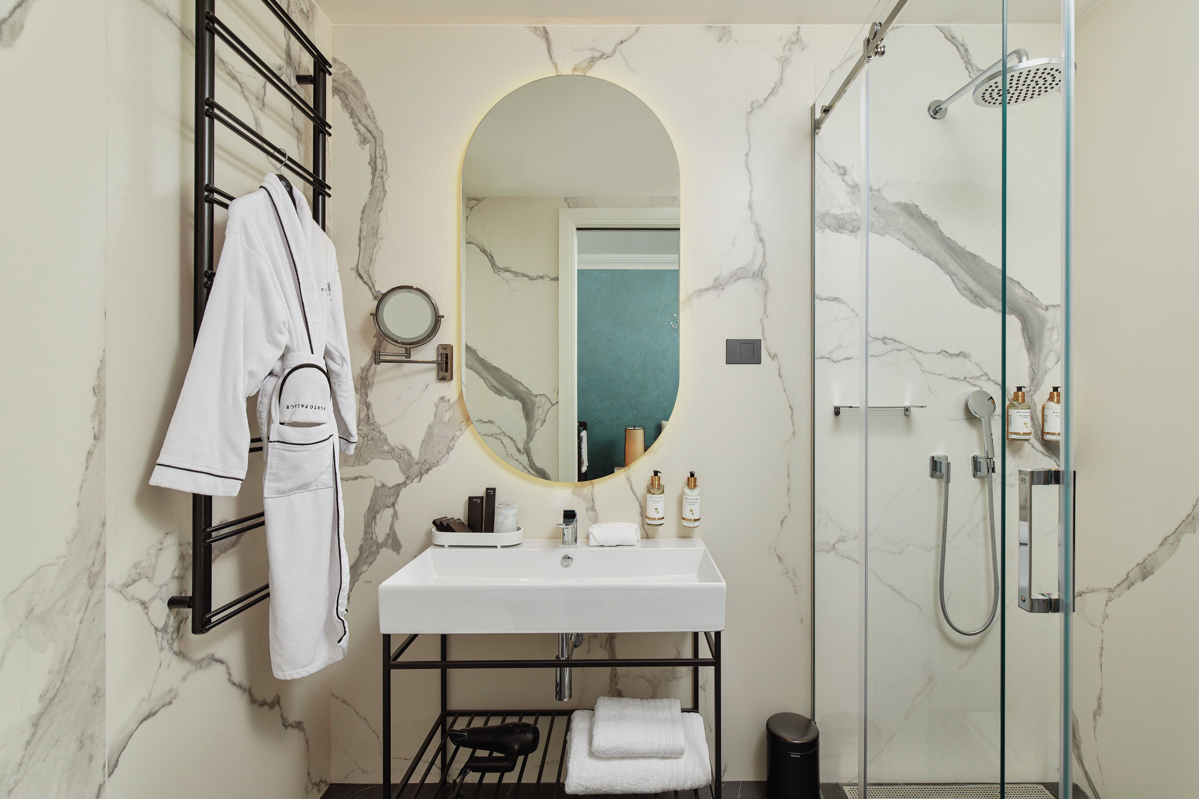 A bathroom with a marble wall, a white sink, a mirror, a glass shower enclosure, and a towel rack.