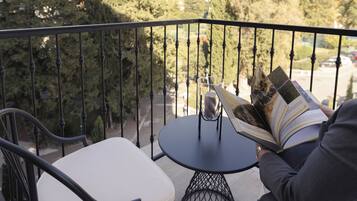 A balcony with a table and chairs, a person reading a book, and a view of trees and buildings in the background.