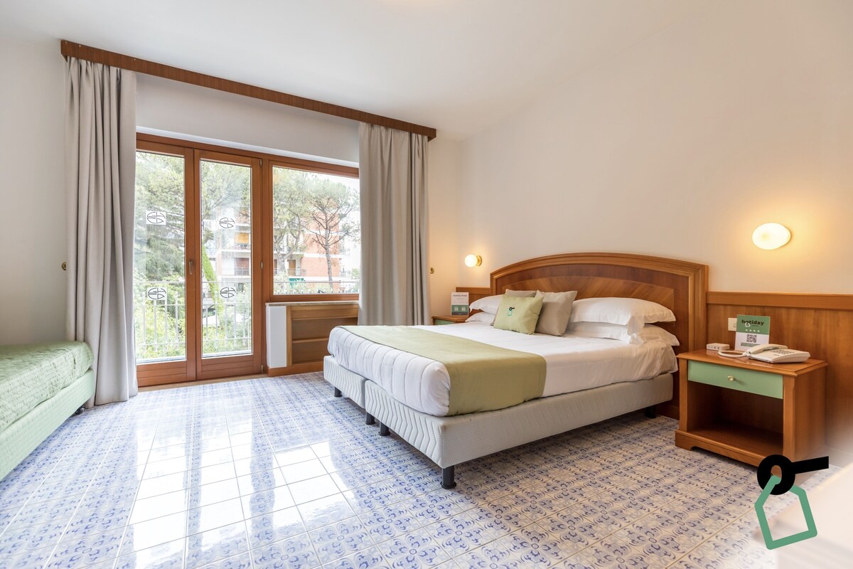 Ein Hotelzimmer mit einem Bett, einem Nachttisch, einem grünen Sofa und einem großen Fenster mit Vorhängen.