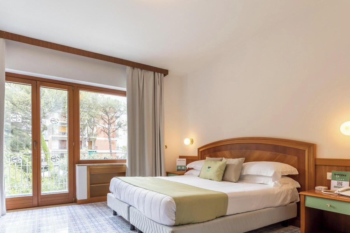 Ein Hotelzimmer mit einem Bett, einem Nachttisch, einem grünen Sofa und einem großen Fenster mit Vorhängen.
