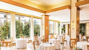 Un restaurante con mesas redondas, sillas y amplios ventanales que ofrecen vistas a árboles y edificios.