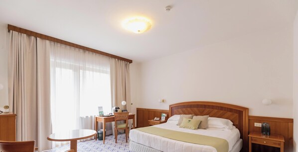 A hotel room with a bed, a desk, a chair, a small table, and a bedside table with a lamp.
