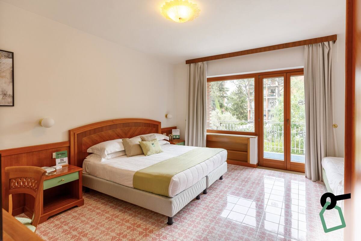 Habitación de hotel con una cama grande, una mesita de noche de madera, un escritorio y una ventana con cortinas.