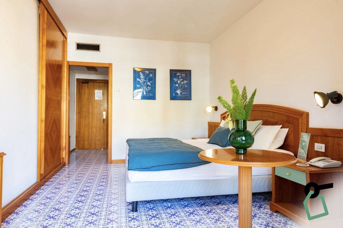 Ein Hotelzimmer mit einem Bett, einem Nachttisch mit einer Vase, einer Lampe und einem Wandtelefon.
