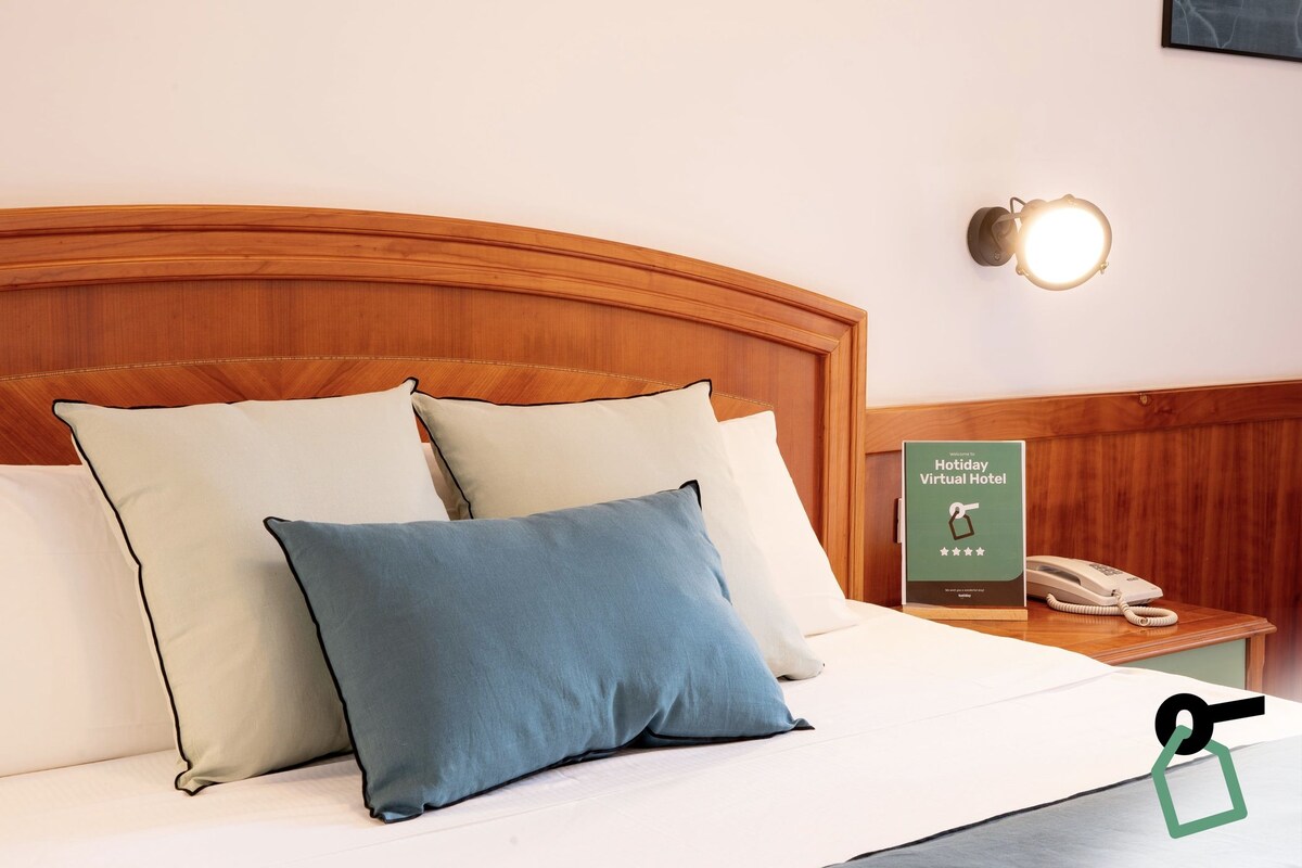 Una cama bien hecha con almohadas, una mesita de noche con un teléfono y un cartel, y una lámpara fijada a la pared.