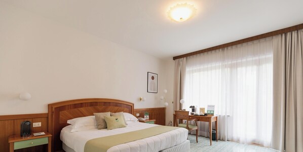 A hotel room with a wooden headboard, a bed with white bedding, a desk with a chair, a nightstand, a bedside table, a lamp, and a window with curtains. - HOTIDAY Room Collection - Sorrento (Sorrento)