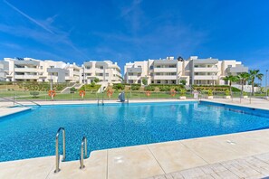 A swimming pool with a clear blue sky, modern white buildings in the background, and a well-maintained landscaped area.