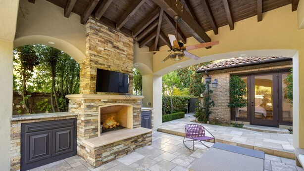An outdoor patio with a stone fireplace, a ceiling fan, and a view of the surrounding greenery.