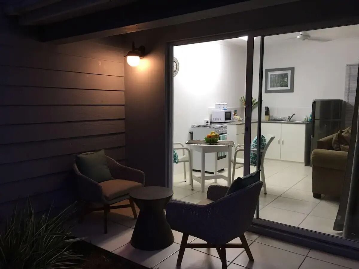 Uma área de pátio com mesa e cadeiras, uma pequena mesa redonda e uma porta de vidro deslizante que dá para uma cozinha e sala de estar.