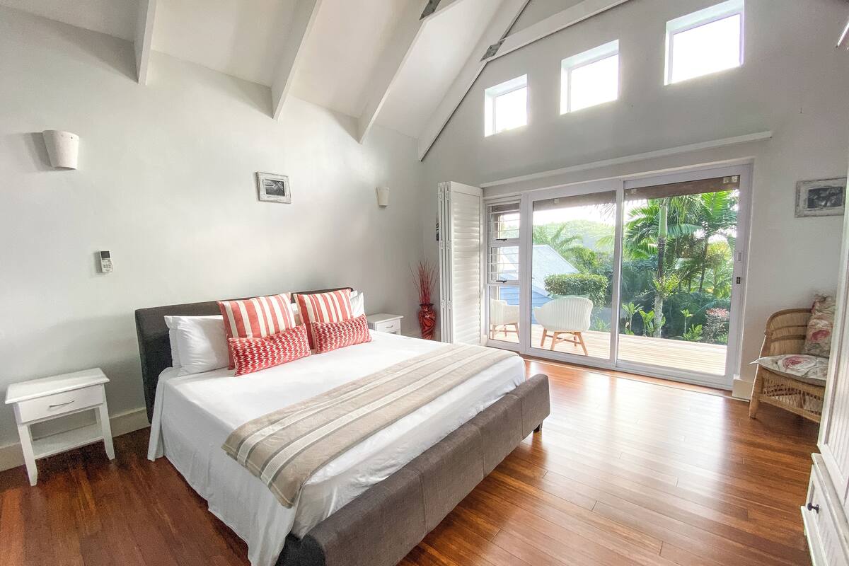 Ein Schlafzimmer mit einem großen Bett, einem Nachttisch, einem Sessel und Blick ins Freie durch eine Schiebetür.