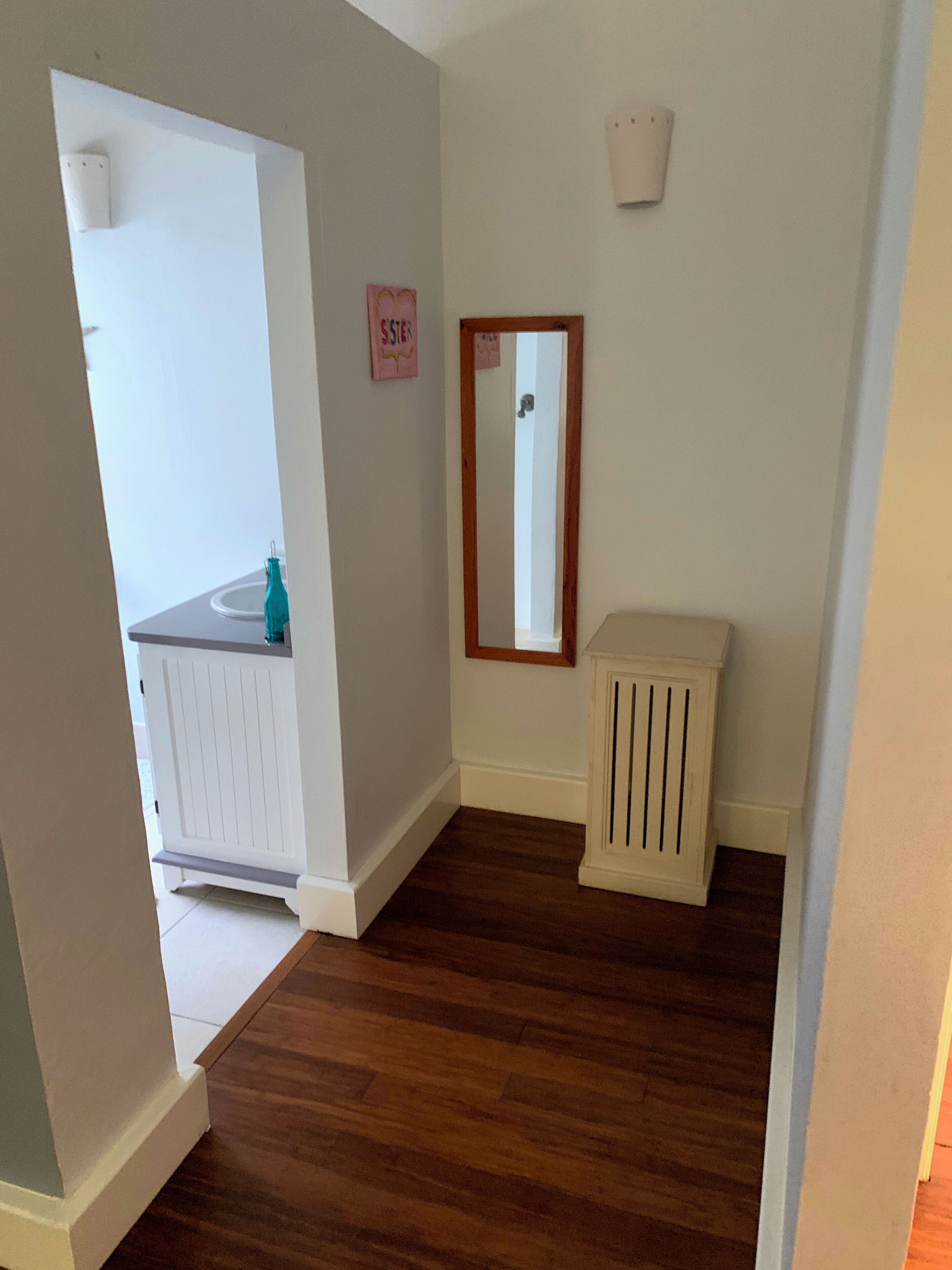 A room with a wooden floor, white walls, a white cabinet, a mirror, and a small sign on the wall.