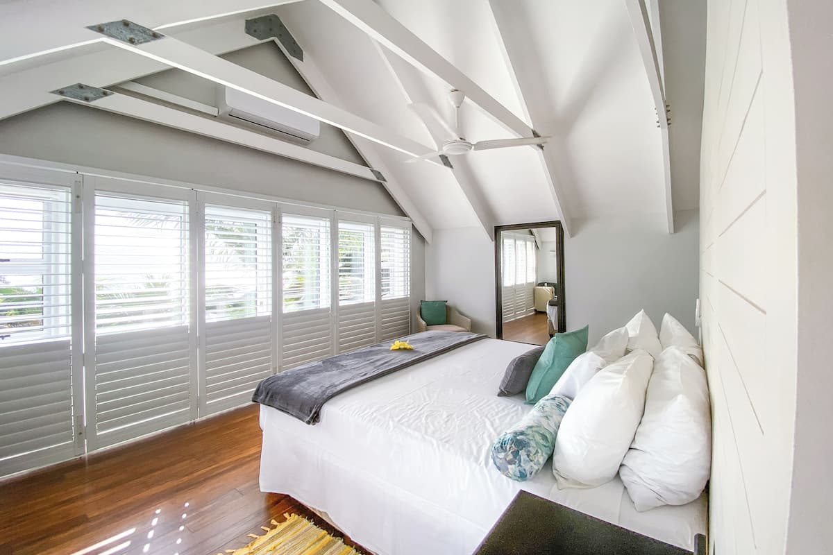 Ein Schlafzimmer mit schrägen Wänden, großen Fenstern mit Jalousien, einem Bett mit weißen und gemusterten Kissen und einem Holzboden.