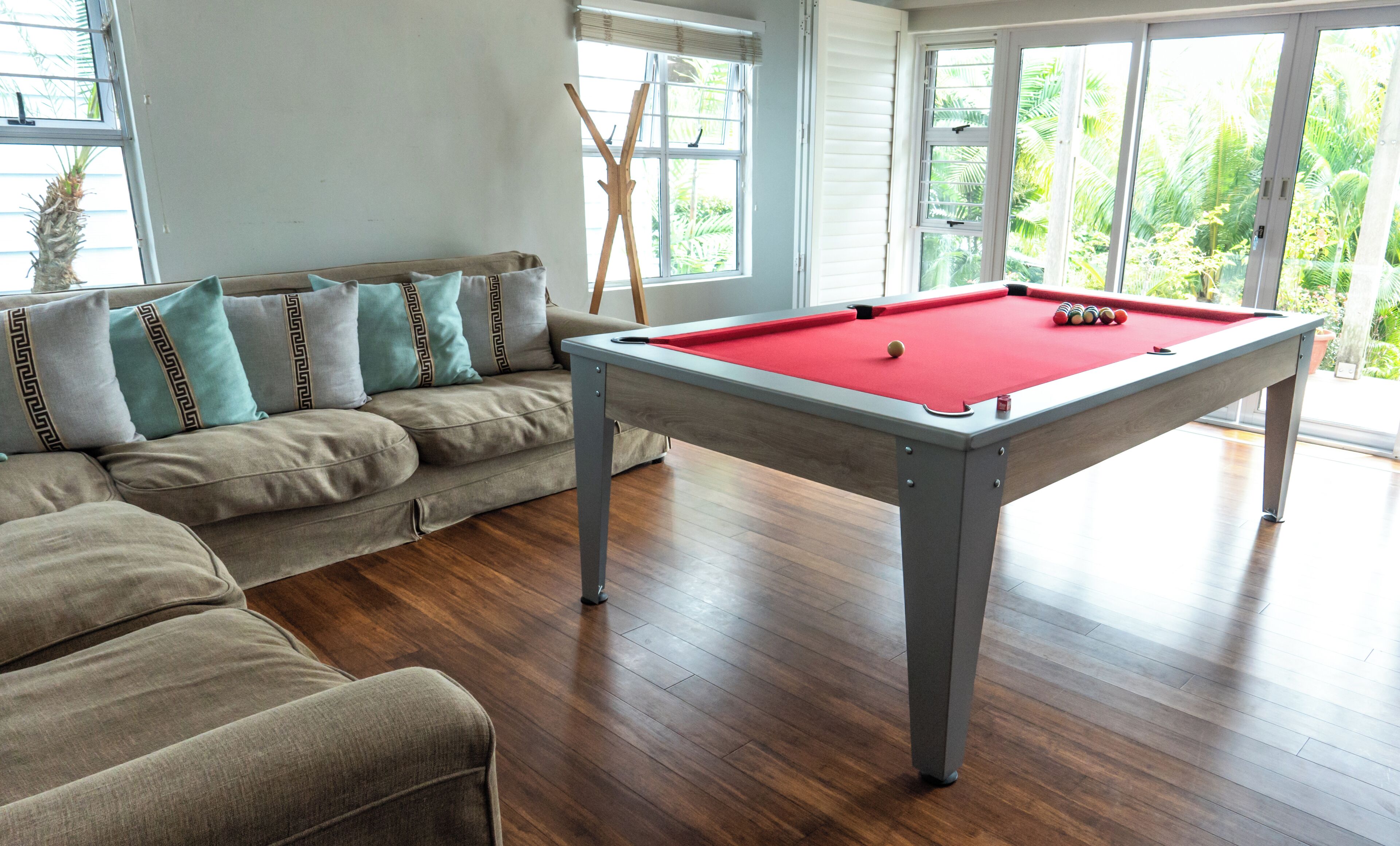 Ein Billardtisch in einem Raum mit Holzboden, einer gepolsterten Couch mit gemusterten Kissen und großen Fenstern mit Blick auf Grünflächen.