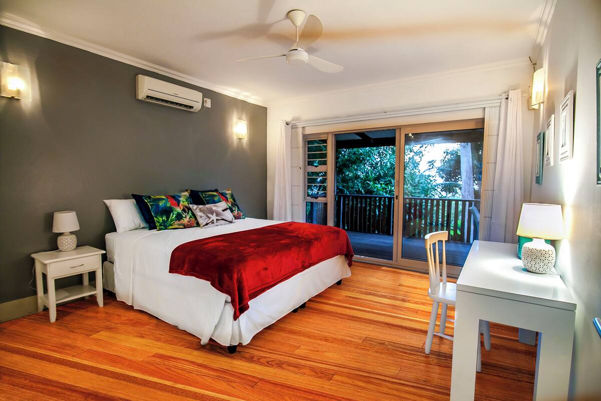 Ein Schlafzimmer mit Bett, Schreibtisch, Stuhl, Deckenventilator und einem Balkon mit Blick auf Bäume.