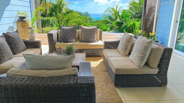 A sunlit outdoor seating area with wicker furniture and cushions, overlooking a tropical landscape and the ocean.