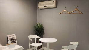 Une chambre d’hôtel moderne, équipée d’un lit, de deux chaises blanches, d’une petite table ronde, d’une plante en pot et d’un climatiseur fixé au mur.