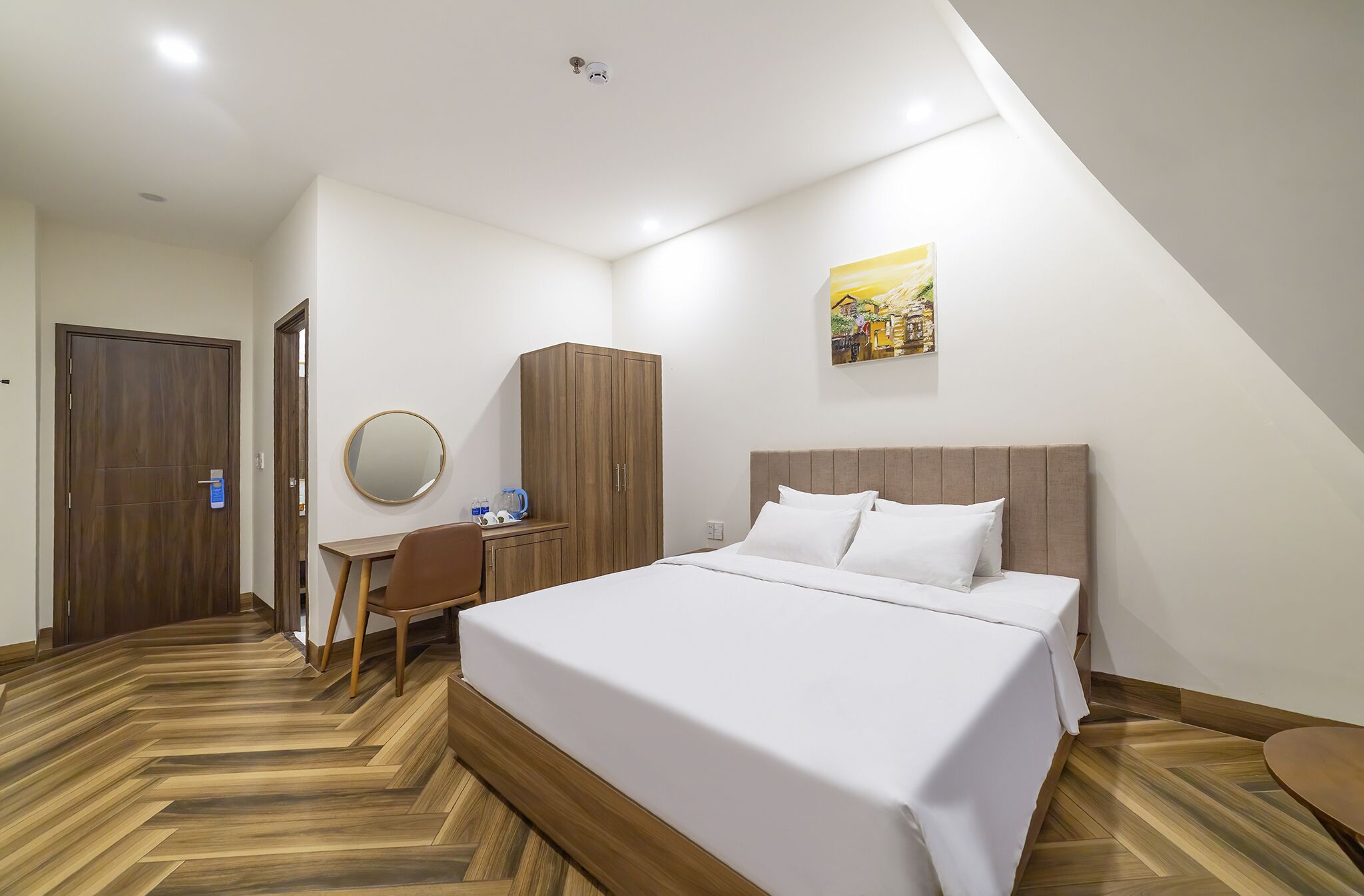 A hotel room with a large bed, a desk with a chair, a wardrobe, and a painting on the wall.